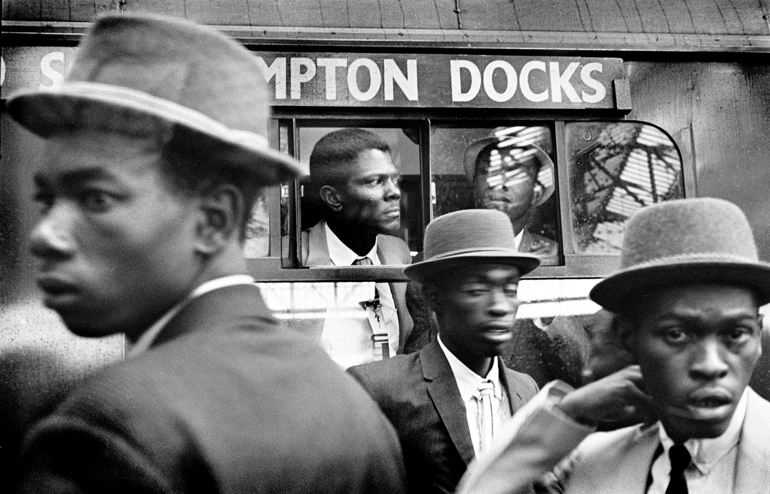 1960s photograph of the Windrush Generation arriving at London Waterloo - men in smart hats in front of a bus.